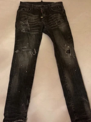 Svarta Dsquared2 jeans med slitningar - Svarta jeans från Dsquared2 i storlek 32 med coola slitningar och distressed detaljer. Byxorna har dragkedja, klassisk femficksdesign och vita färgstänk som ger en edgy vibe. Materialet är jeans och passformen är rak med normal midja.