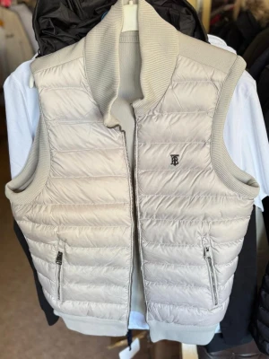 Beige dunväst från Burberry - Snygg beige dunväst från Burberry med quiltad framsida och ribbad krage, axlar och baksida. Västen har två fickor med dragkedja och ett diskret Burberry-emblem på bröstet. Perfekt för lager-på-lager och stilrena outfits.