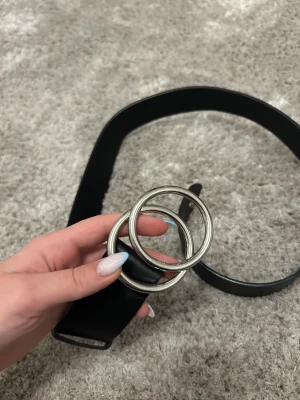 Svart bälte med dubbla ringar - Säljer ett svart bälte med stilrena dubbla silverfärgade ringar som spänne. Bältet är tillverkat i syntetmaterial och har en enkel, klassisk design som passar till jeans eller kjol. Perfekt accessoar för att lyfta din outfit.