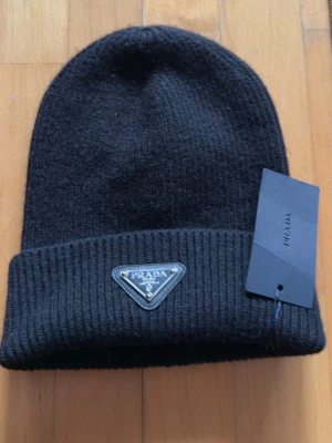 Svart stickad mössa från Prada - Snygg svart mössa från Prada med klassisk ribbstickad design och uppvikt kant. Mössan har en ikonisk metallbricka med Prada-logga framtill och en etikett på insidan. Perfekt för dig som vill ha en stilren accessoar med lyxig känsla.