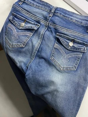 Blå jeans med broderade fickor - Säljer ett par blå jeans med snygga broderade detaljer på bakfickorna och lock med knapp. Jeansen har klassisk femficksdesign, bälteshällor och är tillverkade i slitstarkt denim. Färgen är tvättad blå med ljusare partier för en cool look.