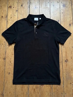 Burberry svart piké - Säljer en svart pikétröja från Burberry med klassisk krage och korta ärmar. Tröjan har knappar framtill och den ikoniska Burberry-rutiga detaljen vid knappslån. Materialet är mjuk bomull och loggan är diskret broderad på bröstet.