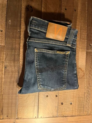 Mörkblå jeans från Nudie Jeans - Jäkligt Snygga mörkblå jeans från Nudie Jeans med klassisk femficksdesign och kontrastsömmar. Jeansen har en rak passform och är tillverkade i slitstarkt denim med läderpatch bak i midjan. Väldigt snygga, rekommenderar!