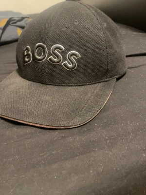 Svart BOSS keps med broderad logga - Säljer en svart keps från BOSS med tydlig broderad logga framtill. Kepsen har klassisk form med böjd skärm och är tillverkad i ett slitstarkt material som känns sportigt och stilrent. Perfekt för dig som vill ha en enkel men snygg accessoar.