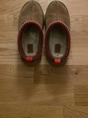 UGG boots med plattform och röd kant - Säljer ett par UGG boots i beige mocka med plattformsula och grovt mönstrad yttersula. Skorna har en stickad kant i rött och vitt samt en dragflik bak. Insidan är fodrad med mjuk, vit päls för extra komfort. Perfekt för kalla dagar och snygg streetstil.