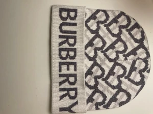 Vit och grå Burberry mössa - Snygg mössa från Burberry i vit och grå färg med stickat mönster av stora B:n och bred uppvikt kant med Burberry-logga. Materialet är stickat och känns mjukt och bekvämt. Perfekt för dig som vill ha en cool accessoar med lyxig känsla.