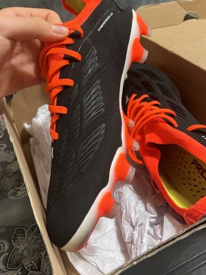 Adidas Predator fotbollsskor svart/orange - Säljer ett par Adidas Predator fotbollsskor med Hybridtouch 2.0-teknologi. Skorna är svarta med vita detaljer och har starkt orange snörning och krage. Insidan är gul med Predator-tryck. Perfekt grepp och komfort för planen.De var rätt store för mig så det är därför jag säljer de annars är de perfekta!.