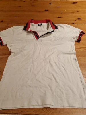 Vit piké från Dolce & Gabbana - Säljer en klassisk vit pikétröja från Dolce & Gabbana med broderad D&G-logga på bröstet. Kragen och ärmsluten har röda och svarta detaljer som ger en sportig vibe. Tillverkad i bomull och har korta ärmar.