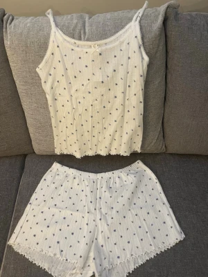 Vit blommig väst från LINDEX - Säljer en söt vit pyjamas set från LINDEX med små blå blommor och diskreta vertikala hålmönster. Toppen har smala axelband och spetskant längs halsringningen och nederkanten. Materialet är mjuk bomullsmix som känns skönt mot huden. Perfekt för dig som älskar skönt och bekvämt i vardagen.