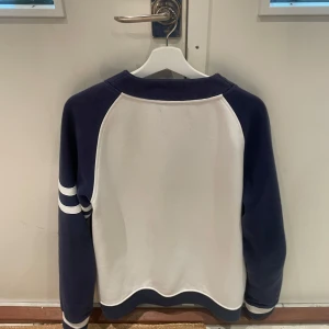 Fet kofta  - Snygg kofta från Bondelid i marinblått och vitt med broderad logga på bröstet och siffran 56. Koftan har långa ärmar med vita ränder, knappar framtill och ribbade muddar. Perfekt cardigan för höst och vinter