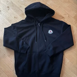 Svart hoodie med dragkedja  - Säljer en stilren svart hoodie med dragkedja och broderad logotyp på bröstet. Tröjan har en justerbar huva med snören och långa ärmar. Perfekt för en avslappnad look.