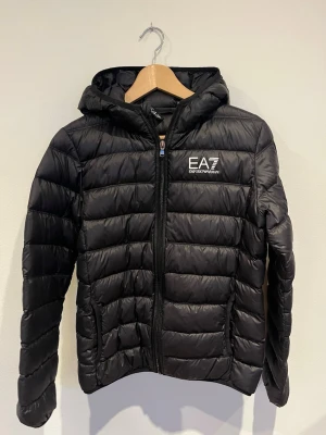 Svart pufferjacka från Emporio Armani - Säljer en svart pufferjacka från Emporio Armani EA7 med huva och dragkedja framtill. Jackan har ett quiltat mönster och logga på bröstet. Perfekt för kyliga dagar och riktigt snygg till streetwear-stilen.