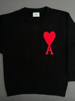 Svart stickad tröja från Ami - Svart stickad tröja från Ami Paris med ett stort rött hjärta och bokstaven A på bröstet. Rund halsringning och ribbade muddar vid ärmslut och nederkant. Perfekt för dig som gillar stilrena detaljer och vill sticka ut med en ikonisk design.