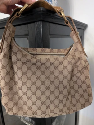 Beige axelväska från Gucci - Säljer en beige axelväska från Gucci med klassiskt GG-mönster i brunt. Väskan har en bred axelrem i brunt skinn med snygga guldfärgade metalldetaljer och dragkedja upptill. Perfekt storlek för det viktigaste och riktigt ikonisk design.