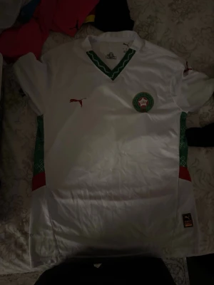 Marocko fotbollströja  - Bilderna visar en vit fotbollströja från Puma med gröna och röda detaljer, samt Marockos landslagsemblem. Ingen sko finns med på bilderna.
