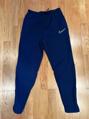 Marinblå Nike dri-fit mjukisbyxor med snörning - Snygga marinblå mjukisbyxor från Nike med klassisk logga på ena benet. Byxorna har elastisk midja med snörning och smal passform som sitter skönt. Perfekta för träning eller chill. Materialet är mjukt och stretchigt för maximal komfort. Storlekslappen saknas men passar typ 160-170 cm. Helt oanvända