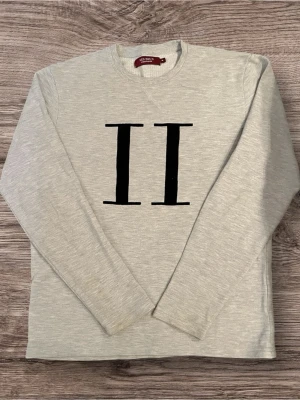 Grå sweatshirt från Les Deux - Snygg grå sweatshirt från Les Deux med stort svart II-tryck på bröstet. Tröjan har rund hals och långa ärmar, perfekt för en chill och stilren look. Materialet är mjukt och känns skönt mot huden. Passar dig som gillar minimalistisk design.