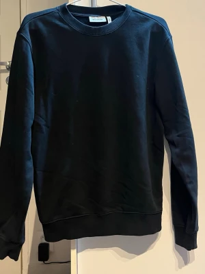 Svart sweatshirt från Weekday XS - En stilren svart sweatshirt från Weekday i storlek XS. Tröjan har rund hals, ribbade muddar vid ärmslut och nederkant samt långärmad passform. Perfekt för dig som gillar enkel och clean stil. Materialet är mjuk bomull som känns skönt mot huden.