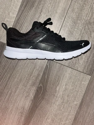Svarta Puma sneakers med vit sula - Snygga svarta Puma sneakers med vit platt sula och SoftFoam komfortinlägg. Ovandelen är i mesh och syntet för bra ventilation och lätt känsla. Skon har snörning och rund tå, perfekt för dig som gillar sportig stil och vill ha bekväma skor till träning.