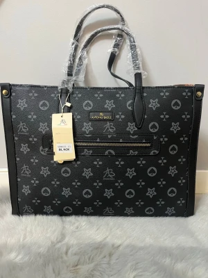 Svart handväska från Antonio Basile - Snygg svart handväska med grått mönster från Antonio Basile. Väskan har rejäla handtag, guldfärgade detaljer och en praktisk dragkedja framtill. Materialet är skinnimitation och designen har ett coolt, grafiskt mönster med stjärnor och symboler. Perfekt för dig som gillar stilrena accessoarer.