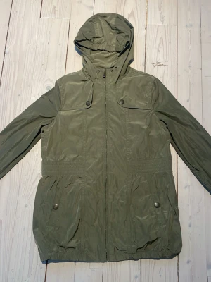 Grön regnjacka från Massimo Dutti - Snygg grön regnjacka med huva från Massimo Dutti. Jackan har dragkedja framtill, stora fickor med knappar och smockdetaljer i midjan för en schysst passform. Perfekt för dig som vill ha en enkel men stilren jacka till vardags.