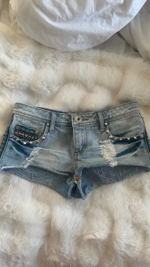 Ljusblå denimshorts med nitar och broderi - Säljer ett par ljusblå denimshorts från True Religion med slitna detaljer, silverfärgade nitar och broderade mönster i turkos och brunt längs fickor och sidor. Shortsen har låg midja och klassisk femficksdesign. Perfekt för dig som gillar edgy och unik stil.