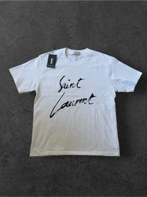 Vit Saint Laurent t-shirt med tryck | M - Säljer en clean vit t-shirt från Saint Laurent med svart handskrivet logotryck på framsidan. Klassisk rund hals och korta ärmar. Materialet är mjuk bomull som känns skönt mot huden. Perfekt för dig som gillar stilrena designer och vill ha något från ett ikoniskt märke.