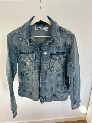 Blå jeansjacka från Zara - Blå jeansjacka från Zara med slitna detaljer och klassisk krage. Jackan har silverfärgade knappar framtill, två bröstfickor med lock och långa ärmar. Stl S.