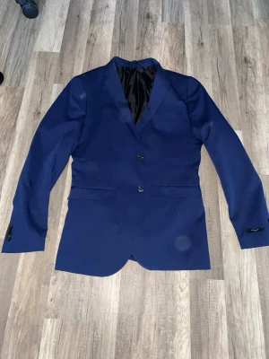 Marinblå kostym med kavaj och byxor - Snygg marinblå kostym med klassisk kavaj och matchande kostymbyxor. Kavajen har två knappar framtill, fickor med lock och knappar vid ärmslut. Byxorna har raka ben och bälteshällor. Materialet känns slätt och har en lätt glansig finish. Perfekt för dig som vill ha en stilren look. Ny pris 1200