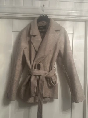 Beige kappa med bälte från Vero Moda - Säljer en snygg beige kappa från Vero Moda i storlek S. Jackan har knytskärp i midjan och långa ärmar. 