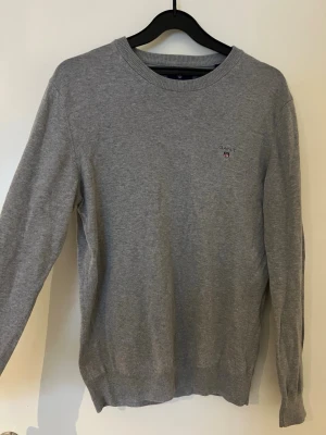 Grå långärmad tröja från GANT - Säljer en stilren grå tröja från GANT med rund hals och diskret logga broderad på bröstet. Tröjan är långärmad och har ribbade muddar vid hals, ärmslut och nederkant. Perfekt för dig som gillar klassisk och enkel design.