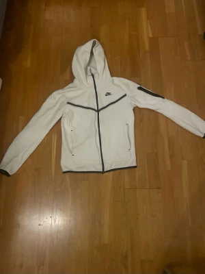 Vitt Nike tech  - Nike vindjacka i vit färg med svarta detaljer och dragkedja hela vägen. Jackan har huva, långärmade ärmar och två fickor framtill. Snyggt svart Nike-logga på bröstet och svarta linjer som ger en sportig look. Perfekt för dig som gillar streetwear.Storlek XS helt ny har inte använt den 