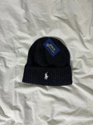 Mörkblå ribbstickad mössa Polo Ralph Lauren - Snygg mörkblå ribbstickad mössa från Polo Ralph Lauren med klassisk broderad logga framtill. Mössan har uppvikt kant och är tillverkad i mjukt material som håller dig varm. Perfekt accessoar för kyliga dagar.