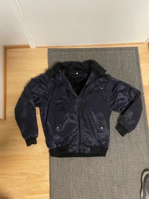 Mörkblå bomberjacka med fuskpäls - Snygg mörkblå bomberjacka med glansig yta och svart fuskpälsfoder. Jackan har flera fickor med dragkedja och tryckknappar, samt ribbade muddar vid ärmslut och nederkant. Perfekt för kalla dagar och har en cool streetstil.