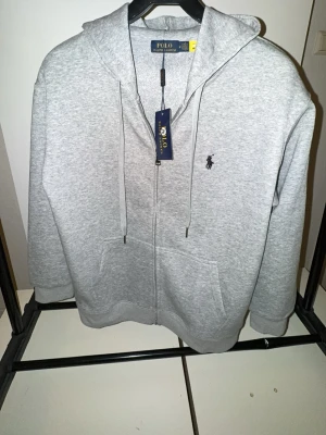 Grå Zip Hoodie - Snygg grå zip hoodie med dragkedja och huva. Klassisk svart logga broderad på bröstet, snörning i huvan och två praktiska fickor framtill. Perfekt för chill dagar och enkel att matcha med jeans eller joggers.