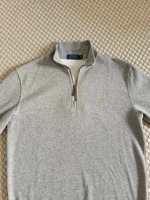 Grå half zip tröja från Polo Ralph Lauren - Snygg grå långärmad tröja från Polo Ralph Lauren med half zip och klassisk krage. Tröjan har ett litet broderat logomärke på bröstet och är tillverkad i mjuk bomull. Perfekt för en chill och stilren look.