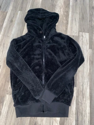 Svart teddyjacka med huva - Mjuk och fluffig svart teddyjacka med huva och hel dragkedja framtill. Jackan har långa ärmar och breda muddar vid ärmslut och nederkant. Perfekt för dig som gillar chill och comfy vibes. Enkel att matcha med jeans eller mjukisbyxor. Märke Amaci and sons. Ny pris 500