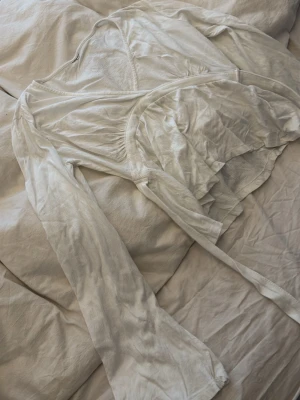 Vit omlottopp med knytband - Säljer en vit omlottopp med långa ärmar och knytband i midjan. Toppen har en v-ringad design och är gjord i ett mjukt, tunt material som känns skönt mot huden. Perfekt att styla med jeans eller kjol för en fräsch look. Har klippt bort lappen då den kliade.