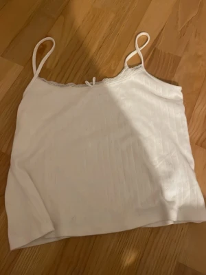 Vitt linne med spets  - söt vit linnetopp från Gina Tricot med tunna axelband, spetskant upptill och en liten rosett i mitten. Toppen har diskret hålmönster och vertikala ränder i tyget. Perfekt för varma dagar och enkel att matcha med jeans eller kjol. Endast använd fåtal gånger och bra skick!