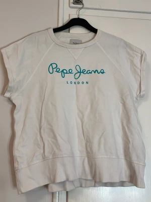 Vit t-shirt från Pepe Jeans - Vit t-shirt från Pepe Jeans med turkost tryck på bröstet. Oversized passform och korta ärmar. Tillverkad i mjuk bomull, perfekt för en avslappnad stil. Klassisk rund halsringning och bredare nederkant. 