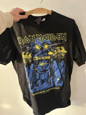 Iron Maiden svart t-shirt - Säljer en svart Iron Maiden t-shirt med fet grafik i blått och gult framtill. Trycket har texten 'Somewhere Back In Time' och coolt motiv med robot och blixtar. Klassisk rund hals och korta ärmar, perfekt för dig som gillar rock och bandmerch.