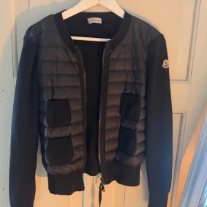 Svart pufferjacka från Moncler - Säljer en svart pufferjacka från Moncler med quiltad front och stickade ärmar. Jackan har rund hals, två ribbade fickor fram och dragkedja. Moncler-logga på ärmen ger extra stilpoäng. Perfekt för dig som vill ha en snygg och bekväm jacka med exklusiv känsla.