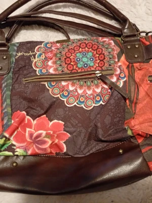 Cool Färgglad handväska från Desigual - Unik handväska från Desigual med blommigt och mandala-inspirerat mönster i rött, brunt, grönt och blått. Väskan har detaljer i brunt skinn, dragkedjor och metallspänne framtill. Perfekt för dig som gillar färg och kreativ design.