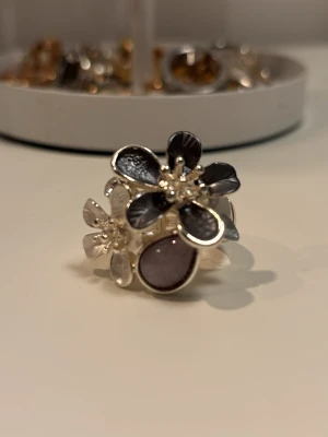 Blommig silverring med gråa detaljer - Snygg och chunky ring i silverfärgad metall med två blommor framtill, en med gråa blad och en med silvriga blad. Blommorna har glittriga detaljer i mitten och ringen har en bred, blank ringbas. Perfekt statement piece för dig som gillar att sticka ut.