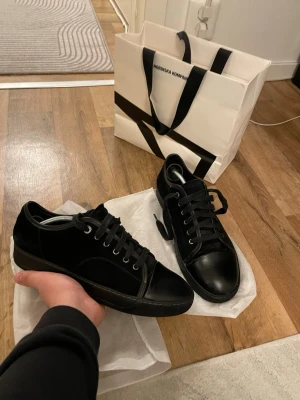 Svarta sneakers från Lanvin i mocka - Säljer ett par svarta Lanvin sneakers med snygg rund tå och platt sula. Skorna har ovandel i mjuk mocka och detaljer i skinn på tå och sidor. Klassisk snörning och diskret design som passar till många stilar. Perfekt för dig som gillar stilrena och exklusiva sneakers.Storlek 42 obs finns ett litet defekt se bild nr5
