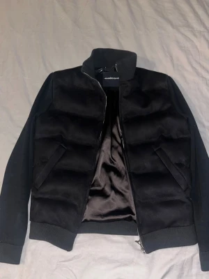 Svart pufferjacka från Manière De Voir - Säljer en svart pufferjacka från Manière De Voir i storlek S. Jackan har quiltad look, hög krage och dragkedja framtill. Materialmix med polyester, elastan och bomull ger en mjuk och skön känsla. Perfekt för dig som vill ha en stilren och modern jacka.