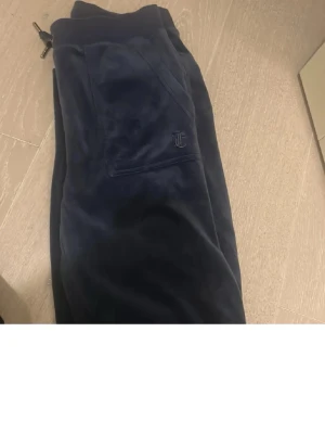 Mörkblå mjukisbyxor från Juicy Couture - Säljer ett par mörkblå mjukisbyxor från Juicy Couture med broderad logga på fickan. Byxorna har snörning i midjan och är gjorda i mjuk och gosig velour som är perfekt för chill dagar. Klassisk design med bekväma fickor.
