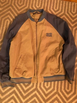 Beige och blå bomberjacka från B.LAKE - Säljer en beige och blå bomberjacka från B.LAKE i storlek M. Jackan har blå ärmar, beige kropp och blått nummer 32 broderat på bröstet. Ribbad krage, muddar och nederkant med blå och beige ränder. Stängs med dragkedja framtill och har två fickor.