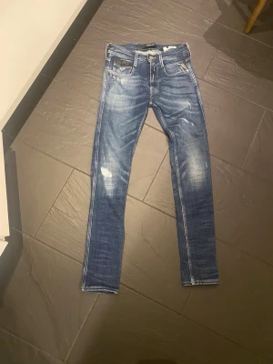 Replay Anbass skinny jeans blå - Säljer ett par Replay Anbass jeans i klassisk blå tvätt med snygga slitningar och skinny passform. Jeansen har fem fickor, dragkedja och knapp, samt detaljerade sömmar på bakfickorna. Perfekt för dig som gillar en smal siluett och cool streetstil.