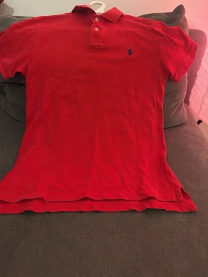 Röd pikétröja Polo Ralph Lauren M - Säljer en röd pikétröja från Polo Ralph Lauren i slim fit-modell. Tröjan har klassisk krage, två knappar vid halsen och den ikoniska broderade loggan på bröstet. Materialet är mjuk bomull och modellen är kortärmad. Perfekt för dig som gillar stilrena och sportiga plagg.